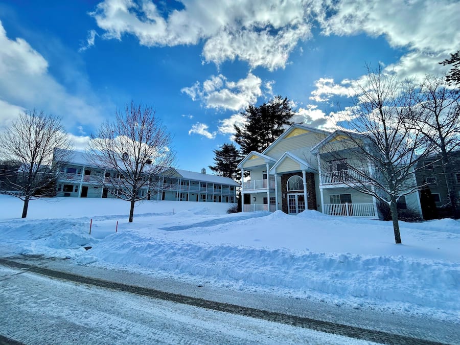 A large, multi-story building with a snow-covered landscape, a clear sky, and bare trees.