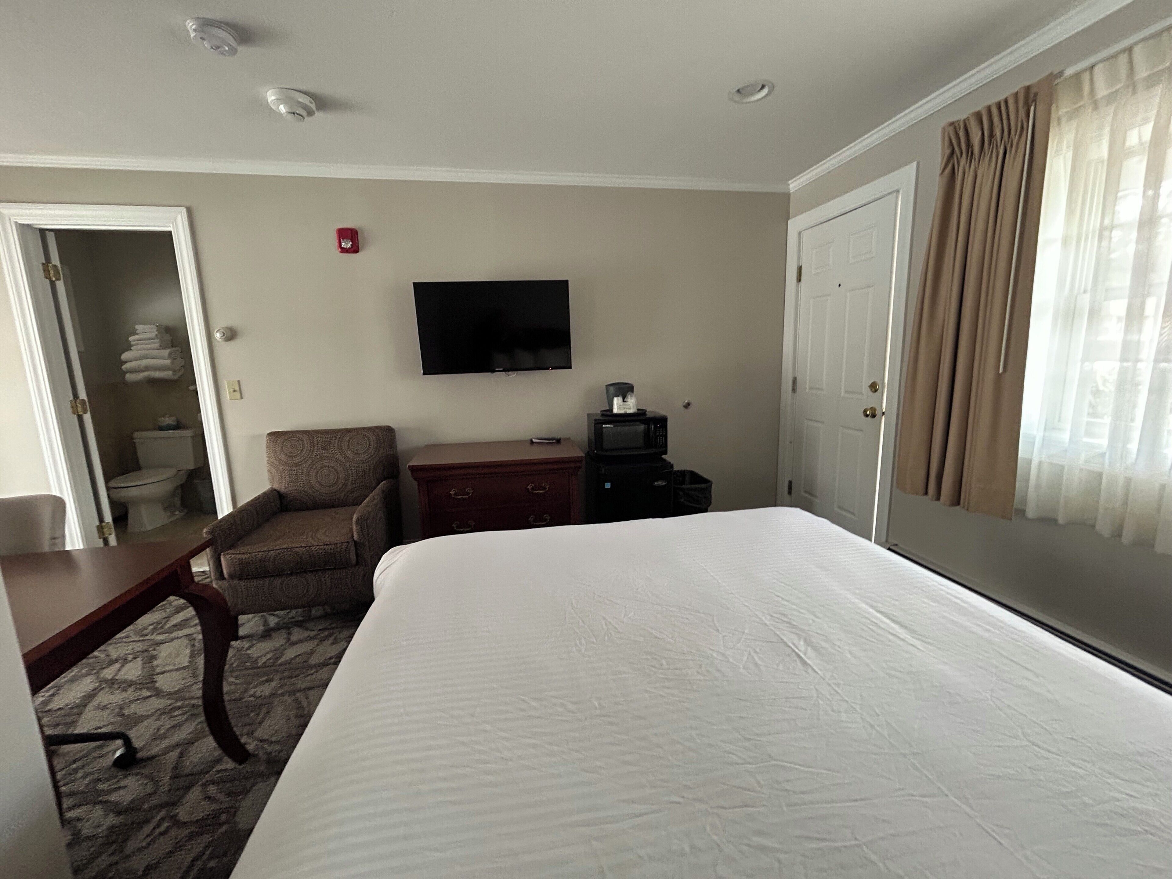 A hotel room with a large bed, a television mounted on the wall, a desk with a chair, and a wardrobe.