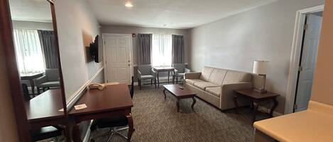 A hotel room with a sofa, a small table, a desk with a chair, a mirror, and a television.