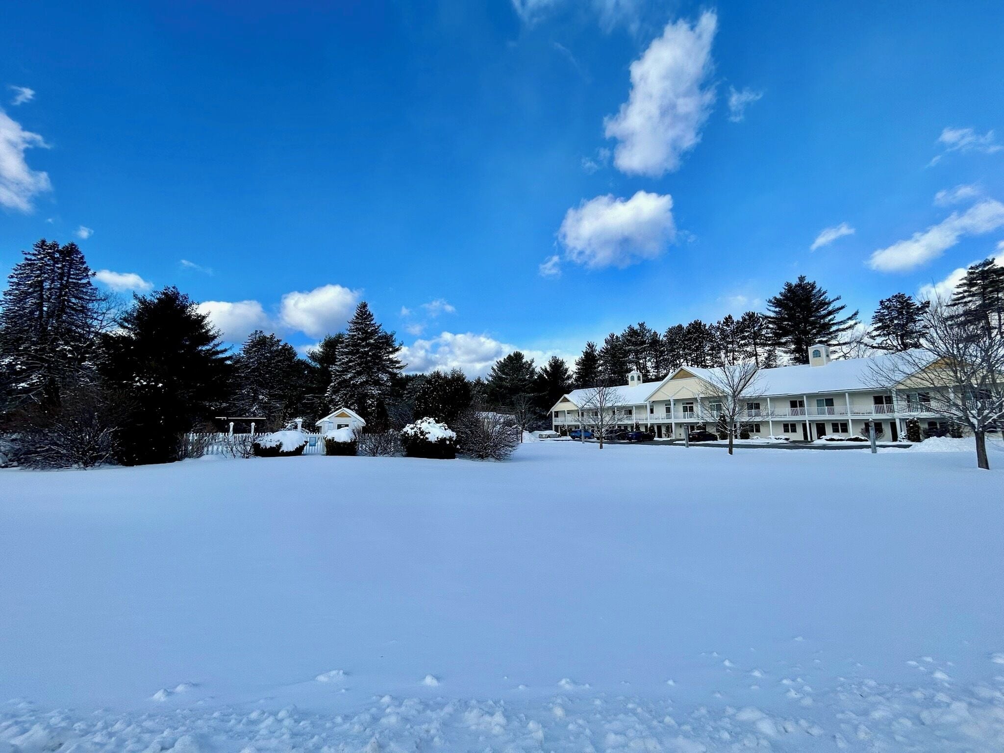 A large, snow-covered house with multiple rooms and a porch, surrounded by trees and a clear blue sky.