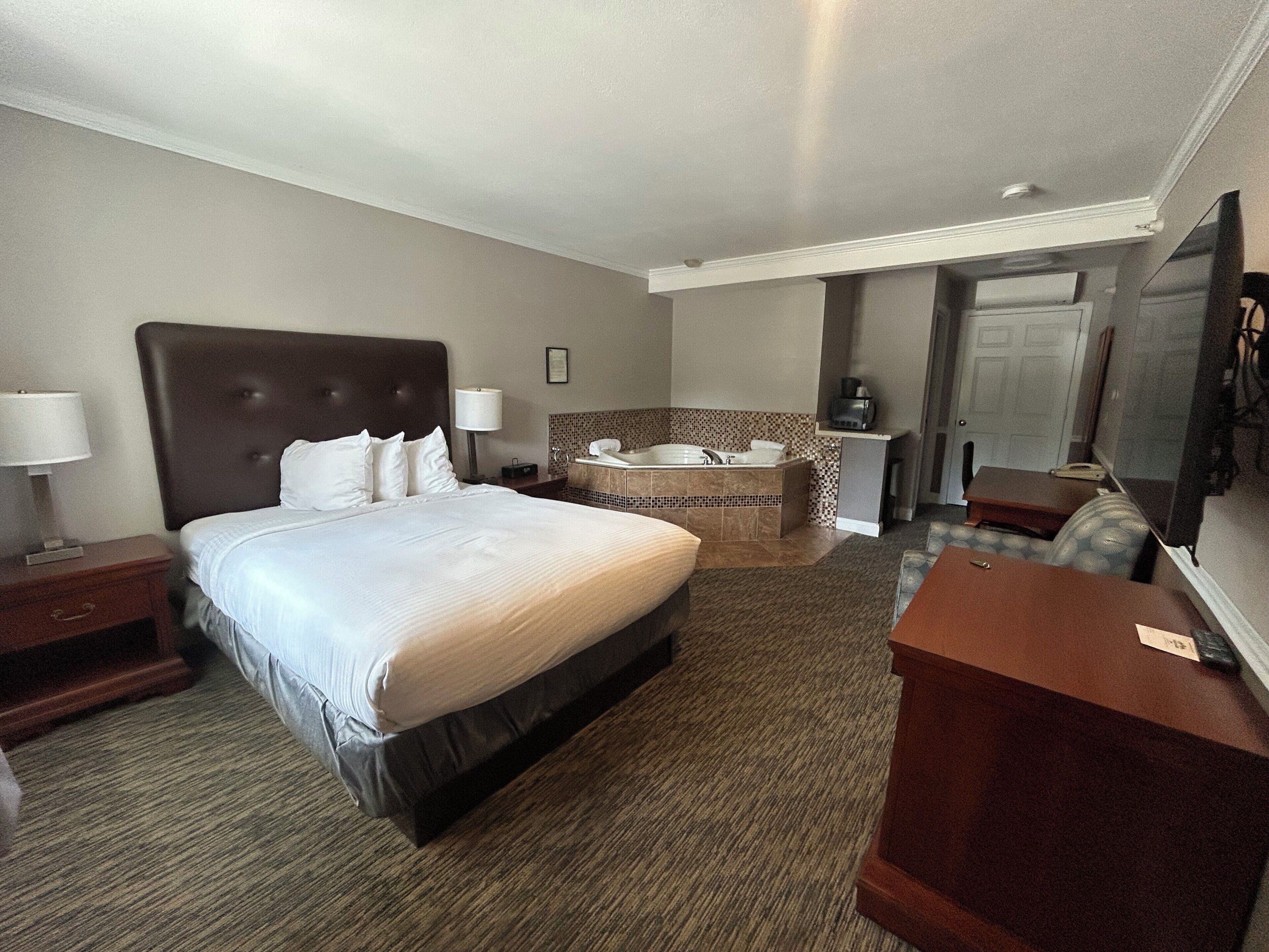 A hotel room with a large bed, a sitting area with a desk, a bathroom with a bathtub, and a television.