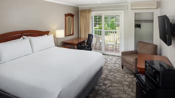 A hotel room with a large bed, a desk with a chair, a television, and a balcony with a view of greenery.