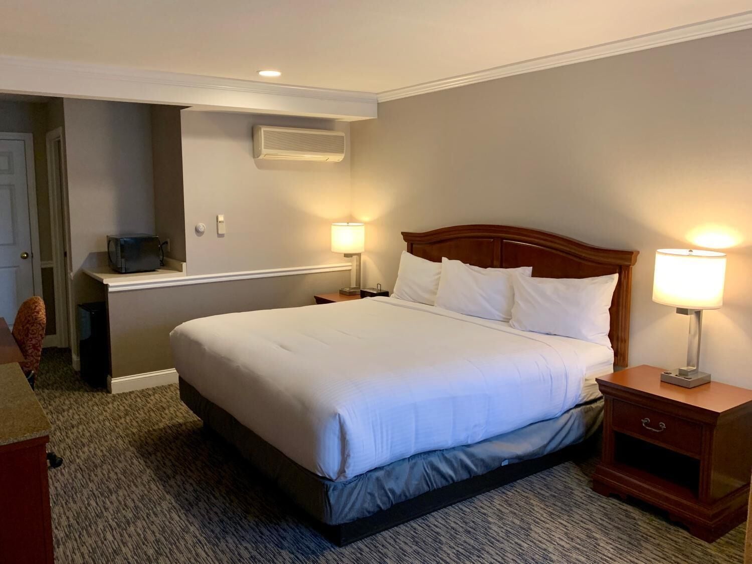 A hotel room with a large bed, two bedside tables with lamps, a desk with a chair, and a wall-mounted air conditioning unit.