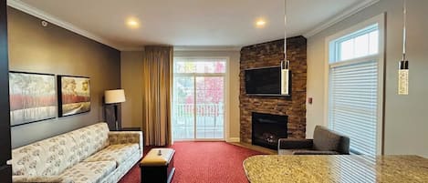 A living room with a sofa, ottoman, and a fireplace. There is a dining table and chairs, and a TV mounted on the wall.