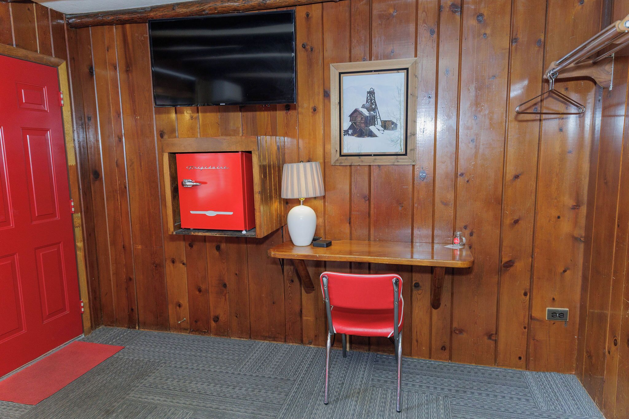 A wooden-paneled room with a red door, a red cabinet, a small table with a lamp, and a red chair.