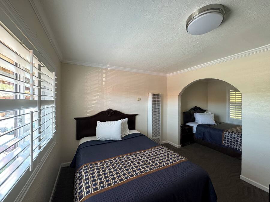 A hotel room with a large bed, a smaller bed, a window with blinds, a ceiling light, and a view of the outside.