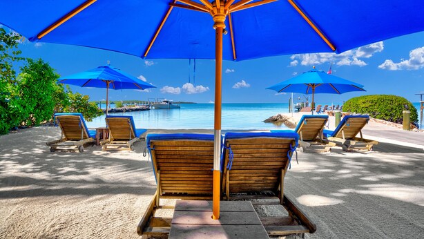 A beachside lounge with wooden lounge chairs and blue umbrellas, overlooking a calm sea and docked boats. - Island Bay Resort (Tavernier)