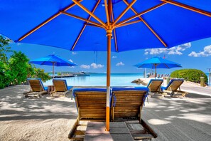 Eine Strandlounge mit Holzliegen und blauen Sonnenschirmen mit Blick auf ein ruhiges Meer und angestellte Boote.