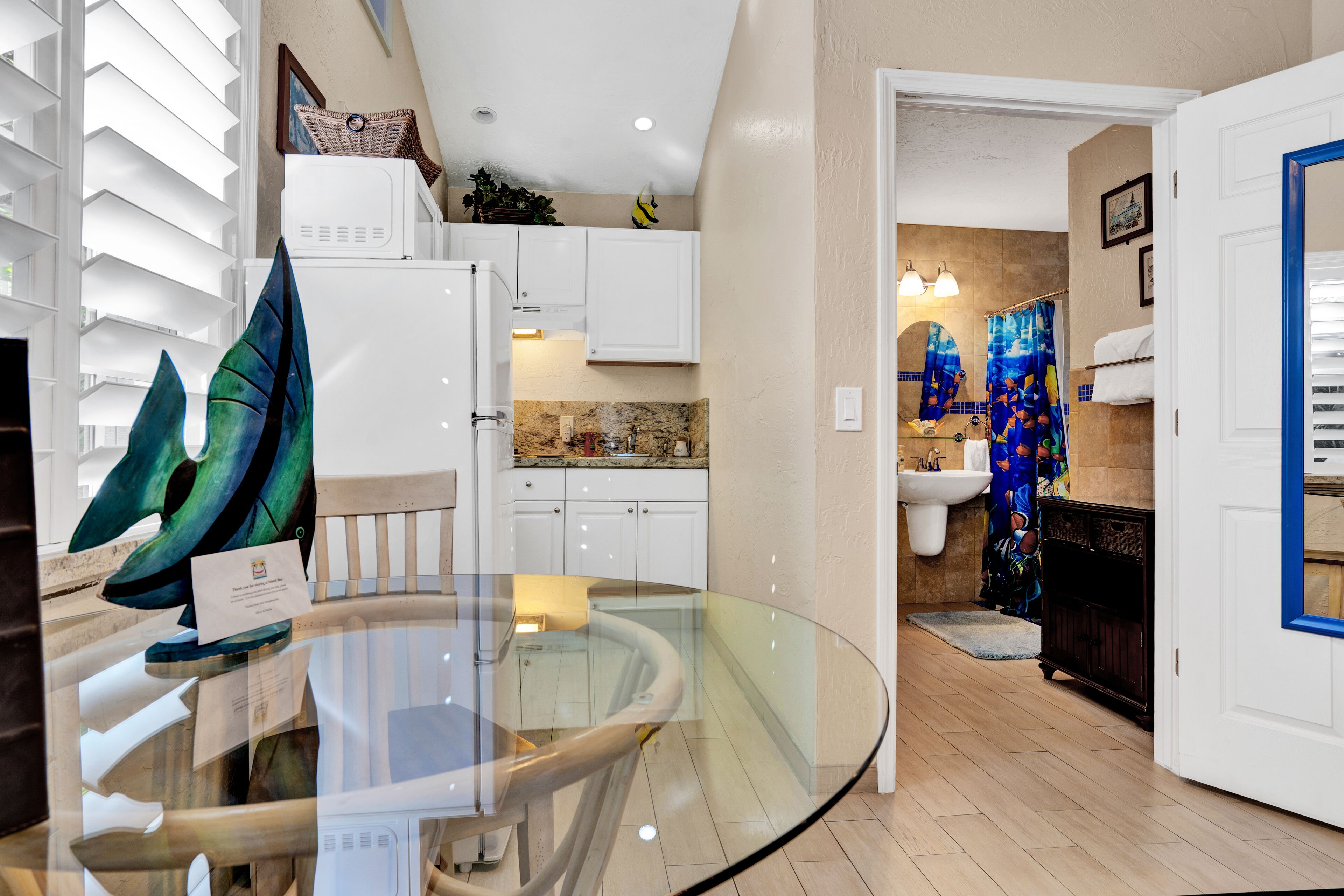 Une cuisine moderne avec des meubles blancs, une table à manger en verre et une vue sur la salle de bain adjacente.
