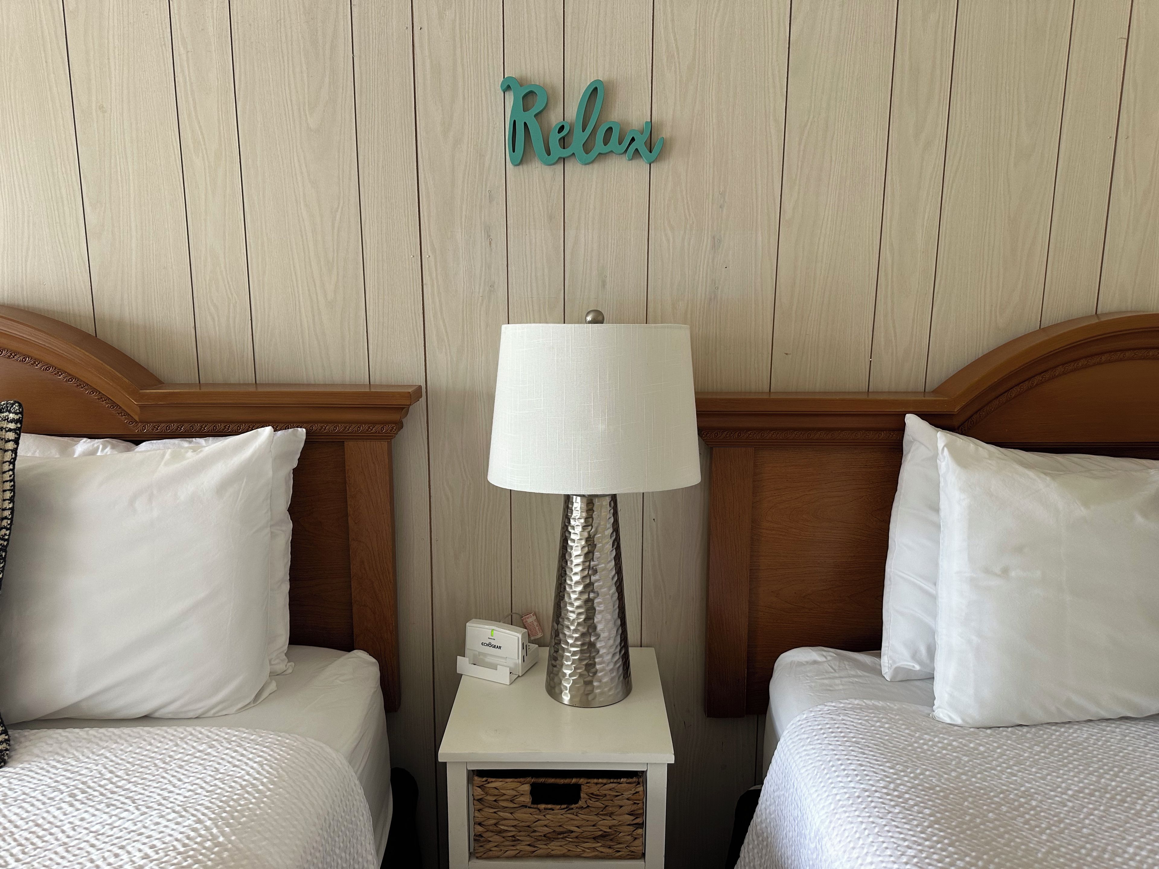 Habitación de hotel con dos camas, una mesita de noche con lámpara, la palabra 'Relax' decorativa en la pared y una pequeña caja sobre la mesita de noche.