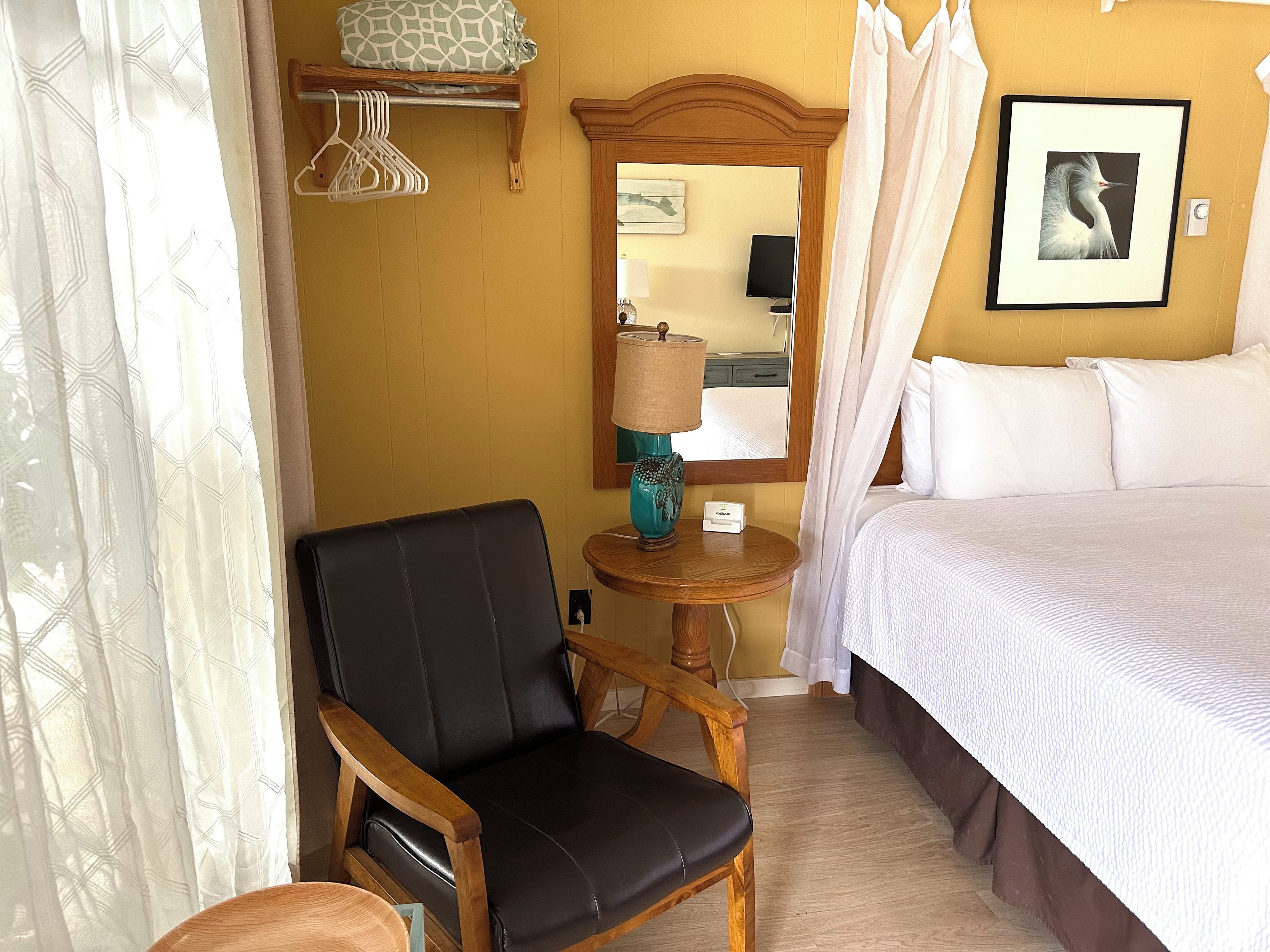 A hotel room with a bed, a chair, a bedside table with a lamp, a mirror, and a picture on the wall.