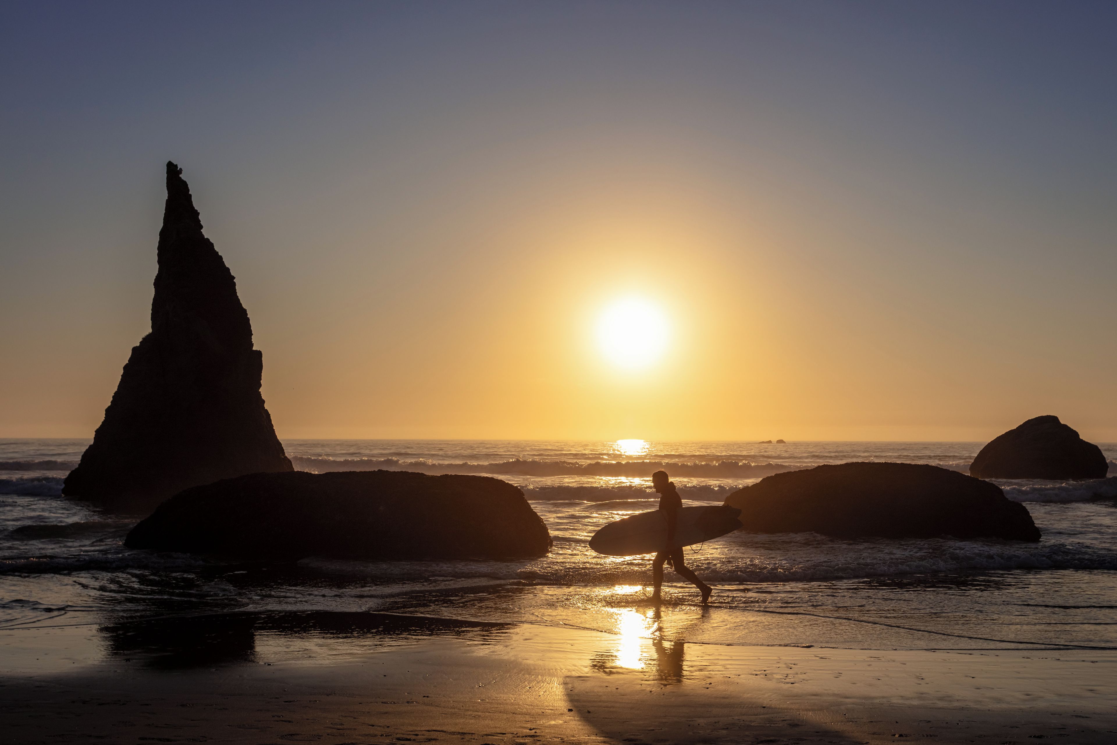 A beach scene with a person carrying a surfboard, large rocks, and the sun setting over the ocean.