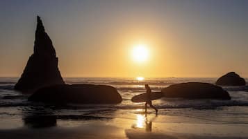 A beach scene with a person carrying a surfboard, large rocks, and the sun setting over the ocean.