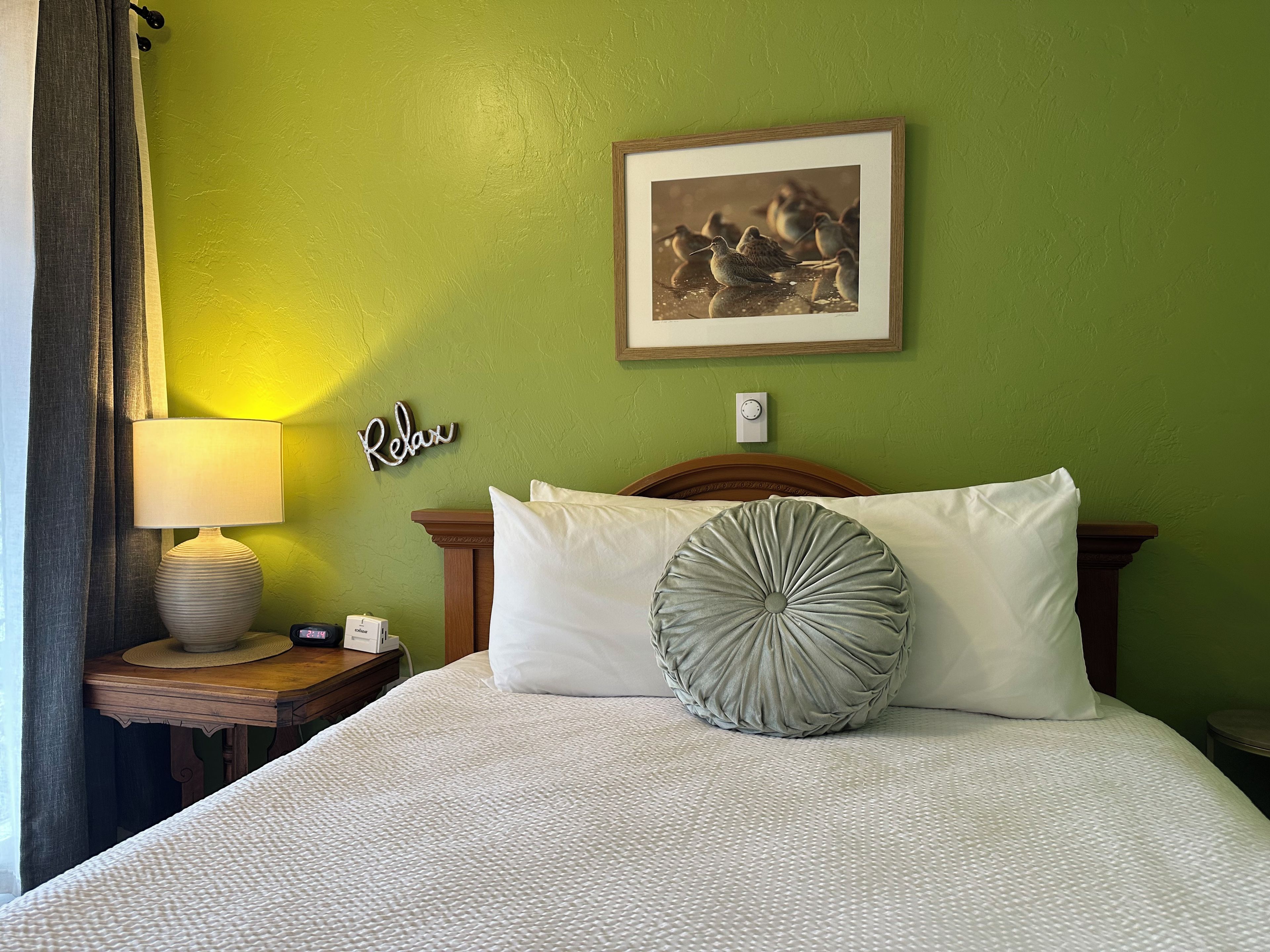 A hotel room with a bed, a nightstand with a lamp, a framed picture on the wall, and a 'Relax' sign.