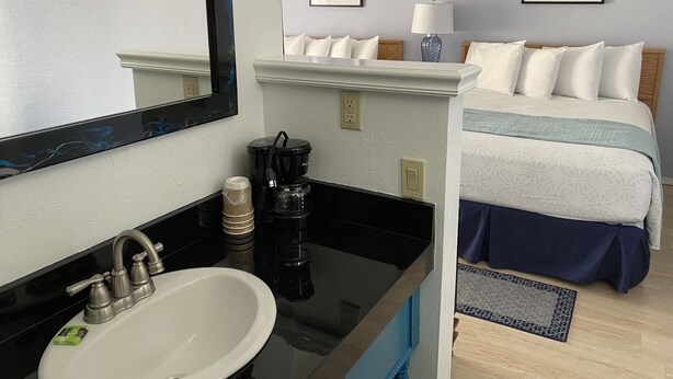 A hotel room with a large mirror, a sink, a coffee maker, a bed with a blue and white bedspread, and two framed pictures on the wall. - La Kris Inn (Bandon)