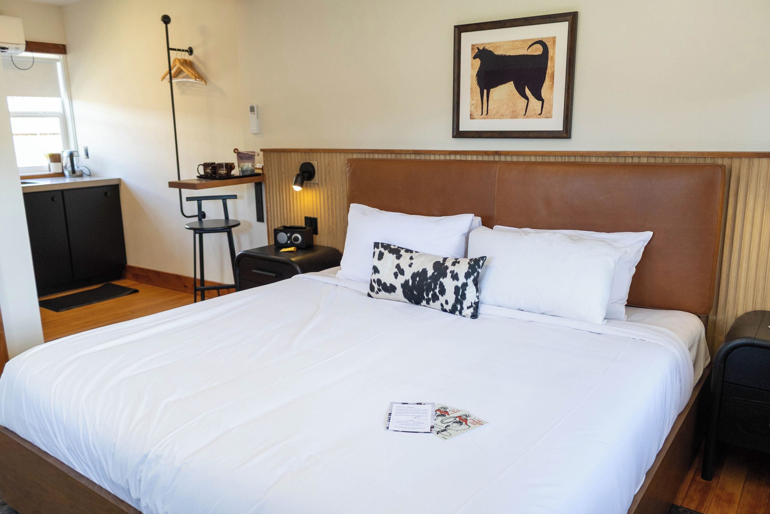 A hotel room with a large bed, a headboard, a desk with a chair, a small table, and a framed picture on the wall.