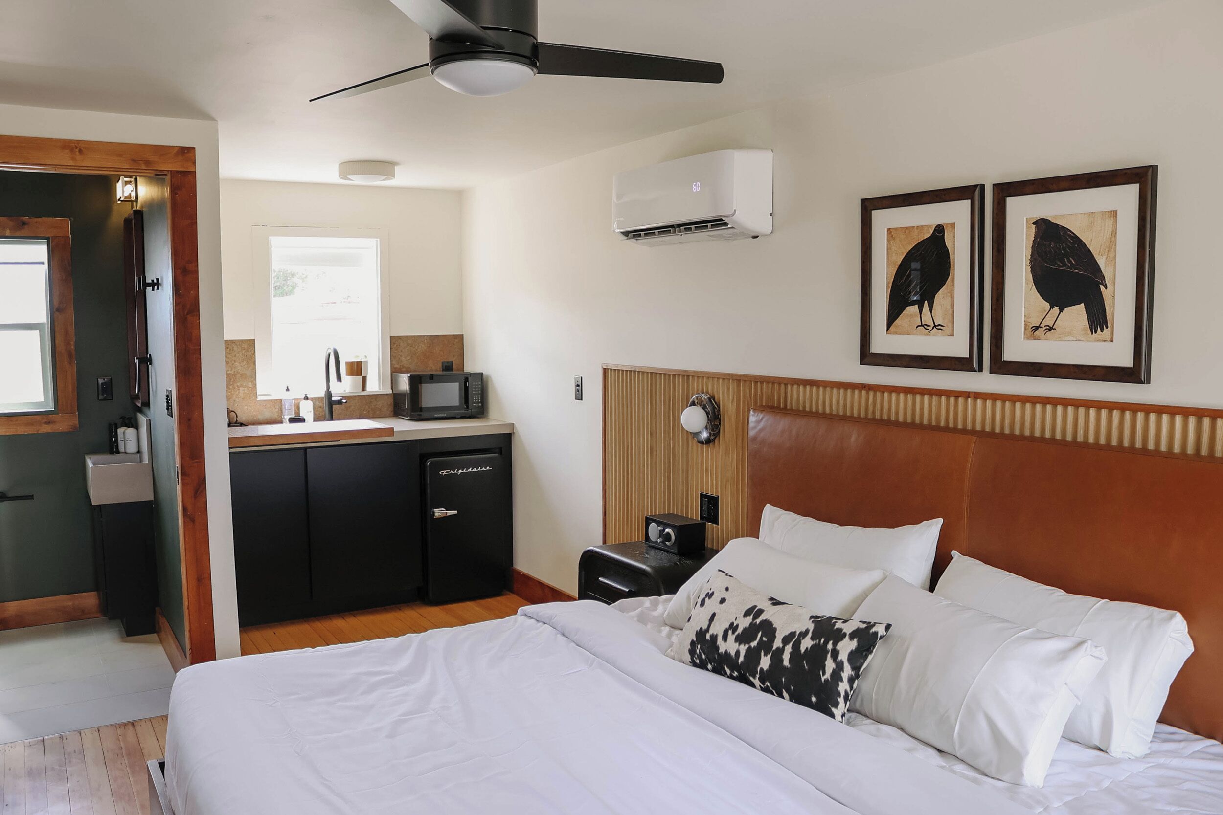 Quarto de hotel com cama, escrivaninha, micro-ondas, mini-geladeira, mesa de cabeceira, relógio e duas fotos emolduradas na parede.
