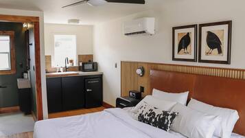 Quarto de hotel com cama, escrivaninha, micro-ondas, mini-geladeira, mesa de cabeceira, relógio e duas fotos emolduradas na parede.