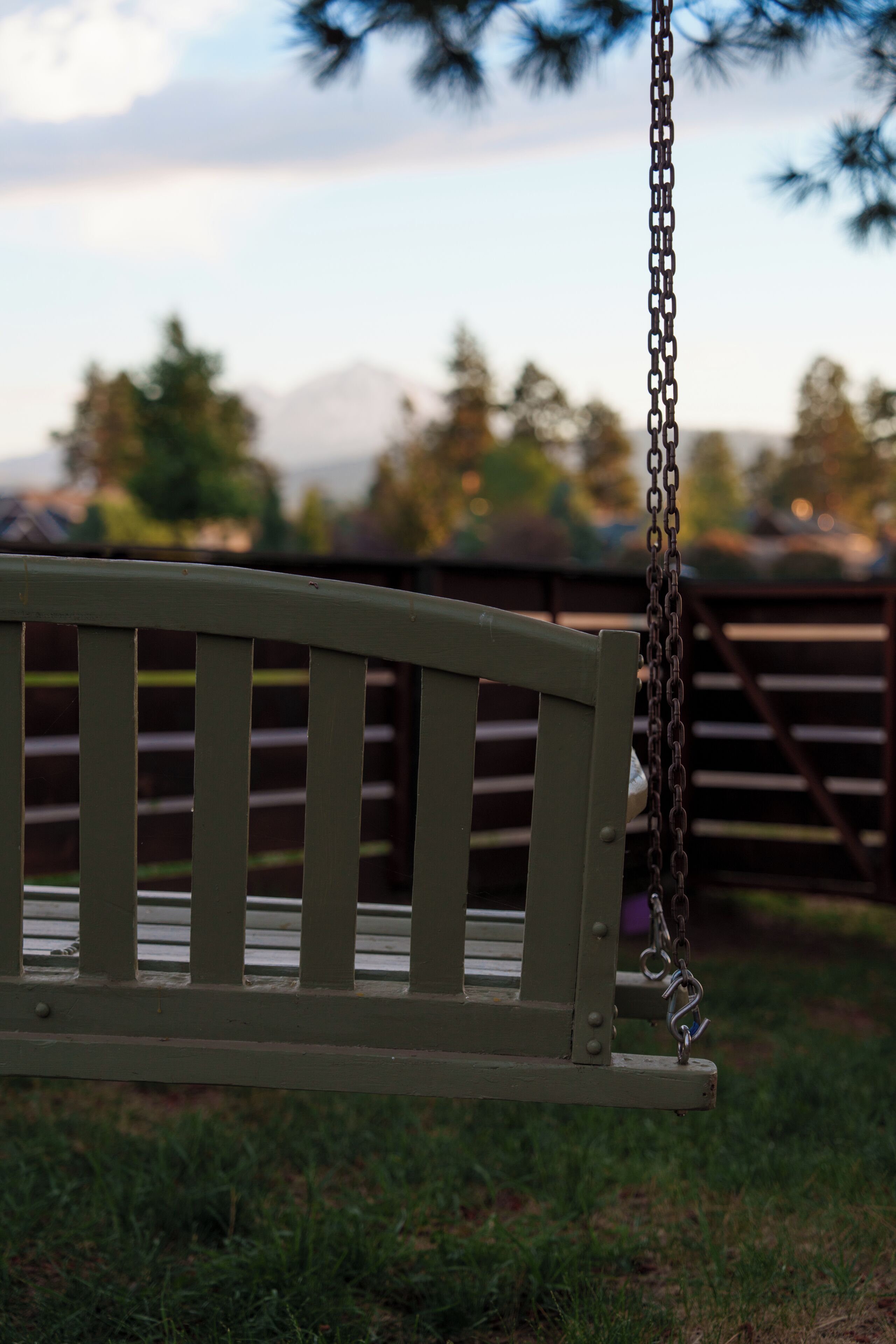 A wooden bench with a metal chain, set against a blurred background of trees and a fence.