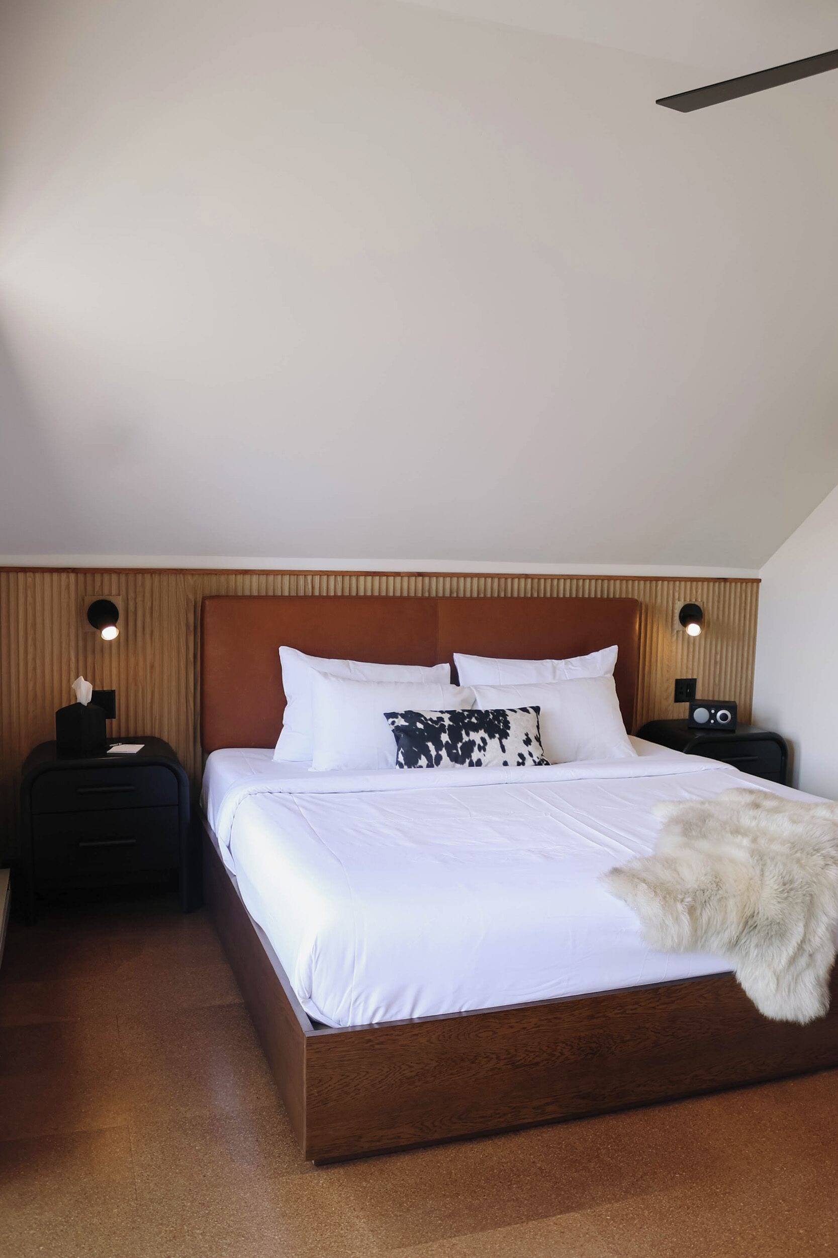 Ein Hotelzimmer mit einem hölzernen Kopfteil, einem großen Bett mit weißer Bettwäsche, zwei Nachttischen mit Lampen und einer Felldecke auf dem Bett.