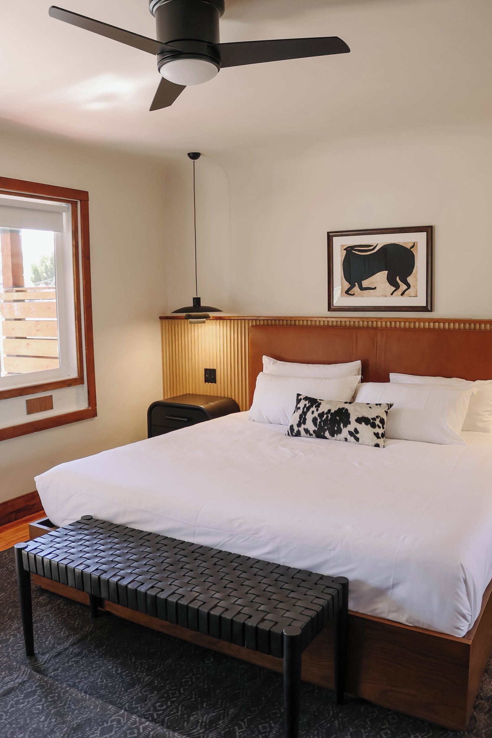 Habitación de hotel con cama, ventilador de techo, mesita de noche, lámpara y un cuadro en la pared.