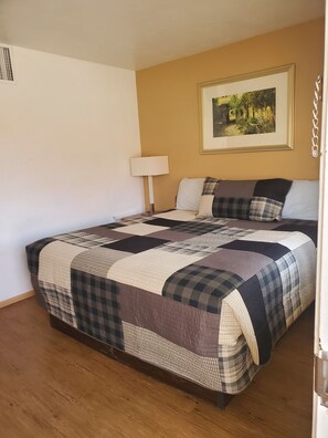 Une chambre avec un grand lit, une table de chevet avec une lampe, un tableau encadré au mur et un plancher en bois.