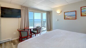 A hotel room with a large bed, a TV mounted on the wall, a small table with chairs, and a balcony with a view of the sea.