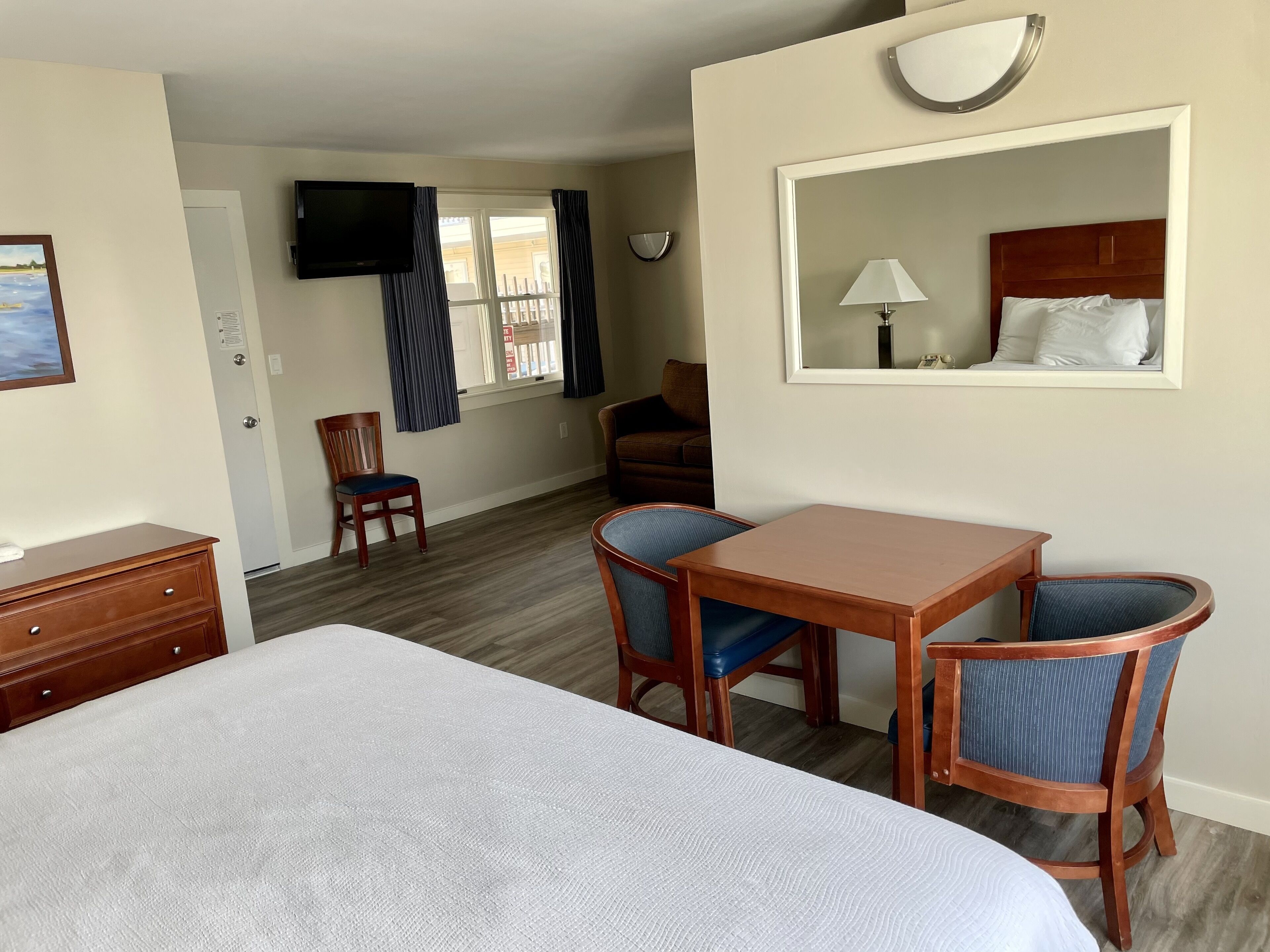 Habitación de hotel con cama, escritorio, dos sillas, televisor, ventana con cortinas y espejo.