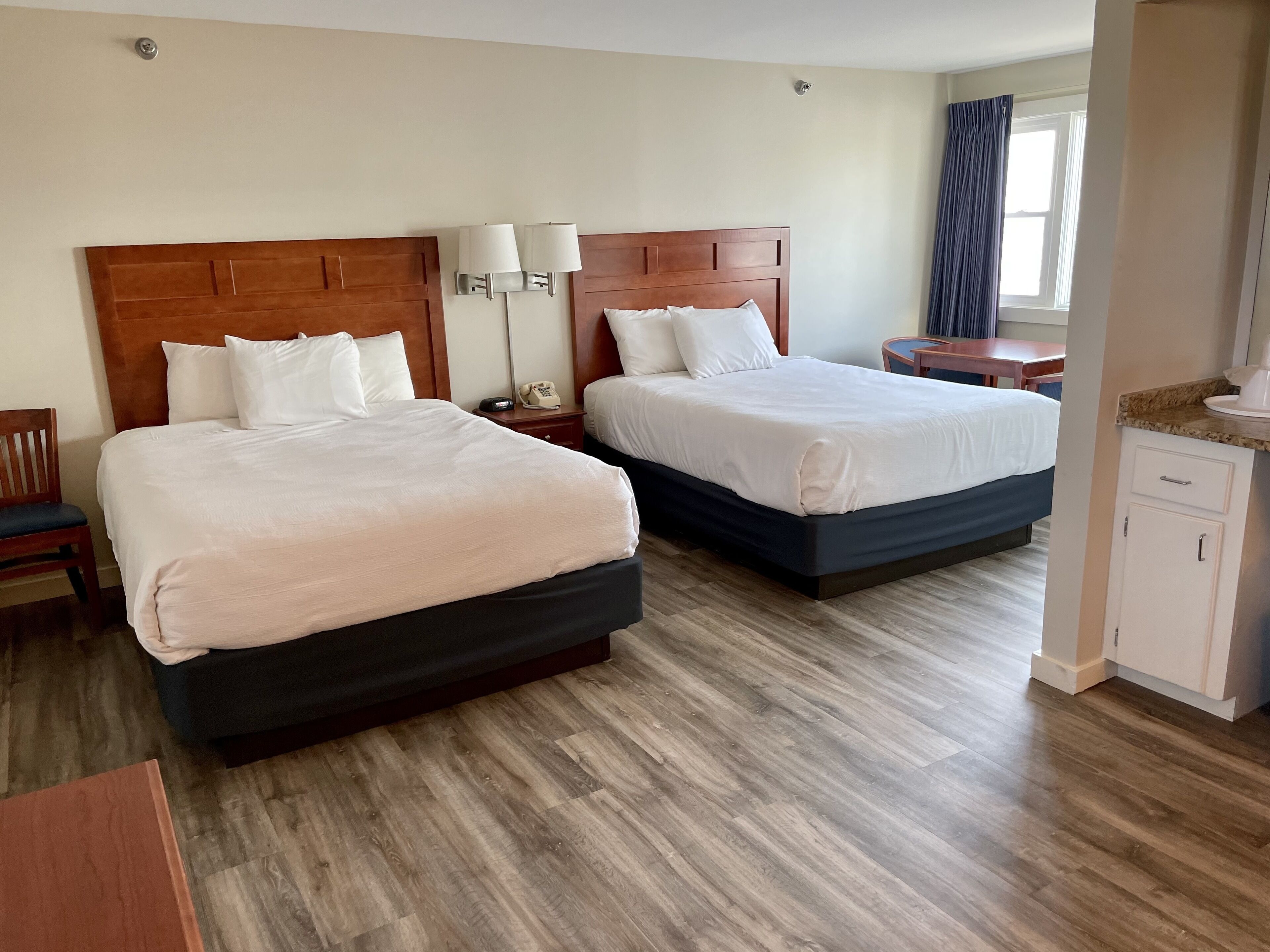 Une chambre d’hôtel avec deux lits, des têtes de lit en bois, du linge de lit blanc, une table de chevet avec un téléphone et un petit bureau avec une lampe.