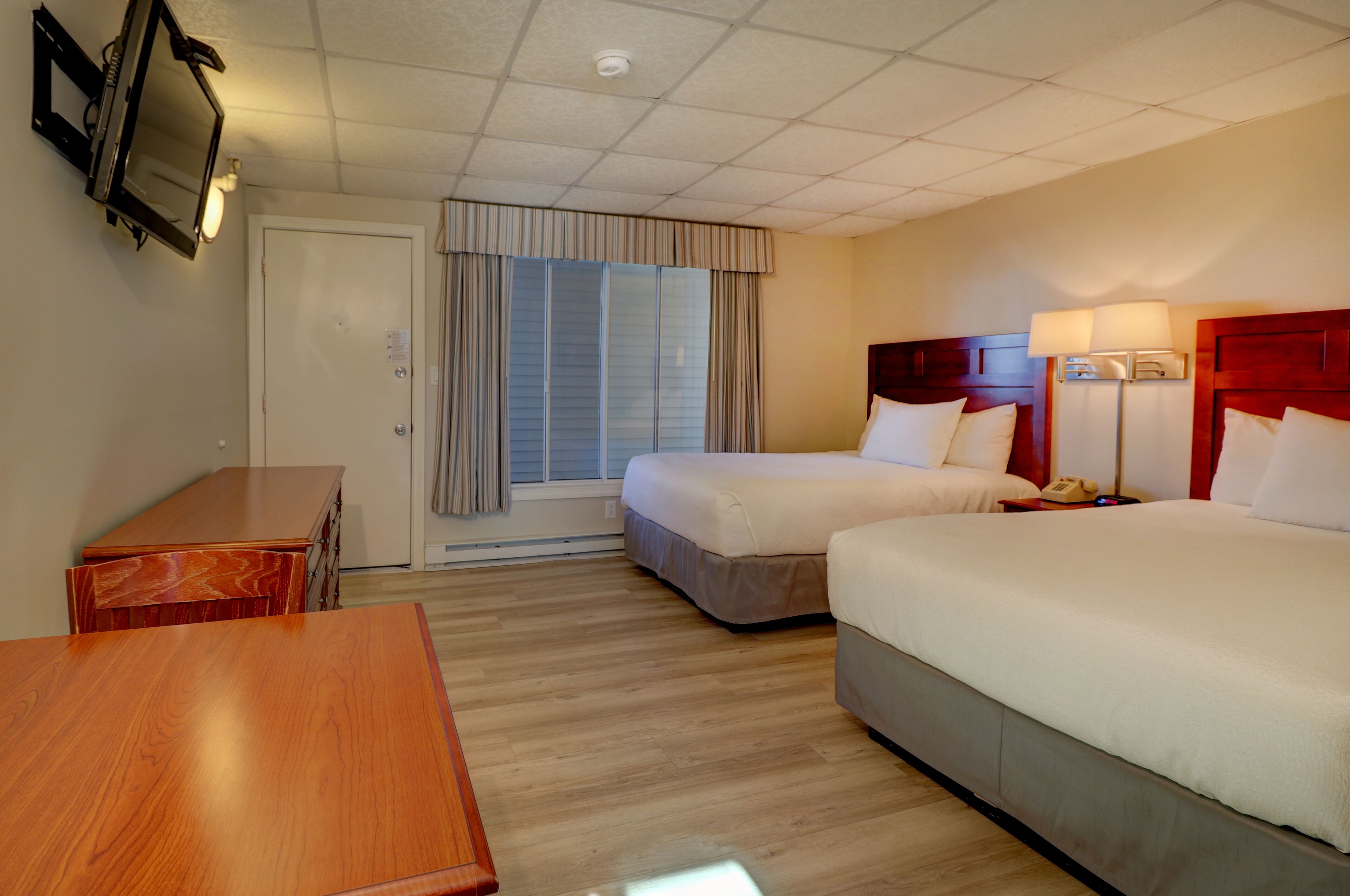 Quarto de hotel com duas camas, uma escrivaninha de madeira, televisão e janela com cortinas.
