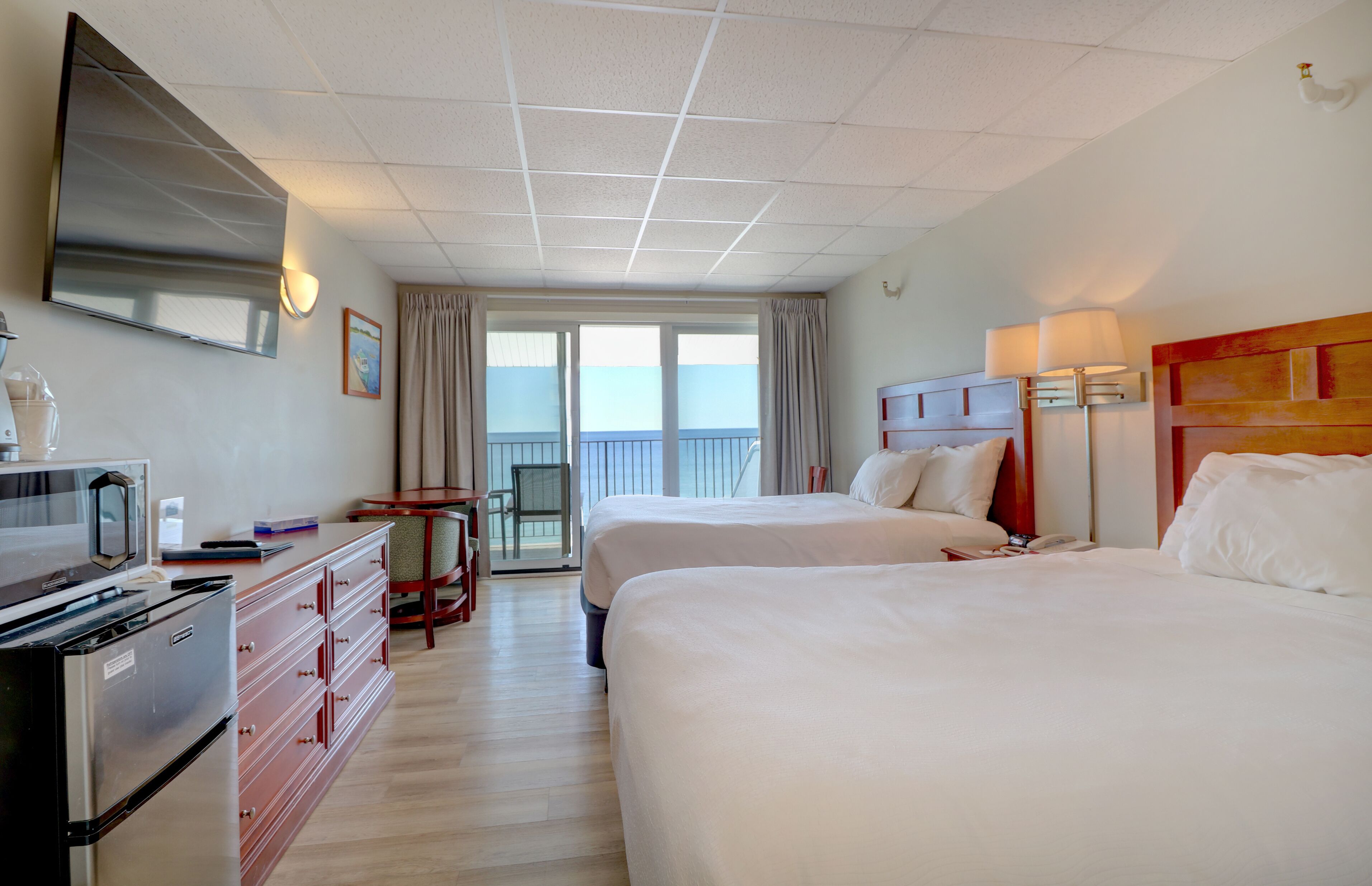 A hotel room with two beds, a TV, a desk with a chair, a minibar, and a view of the balcony.