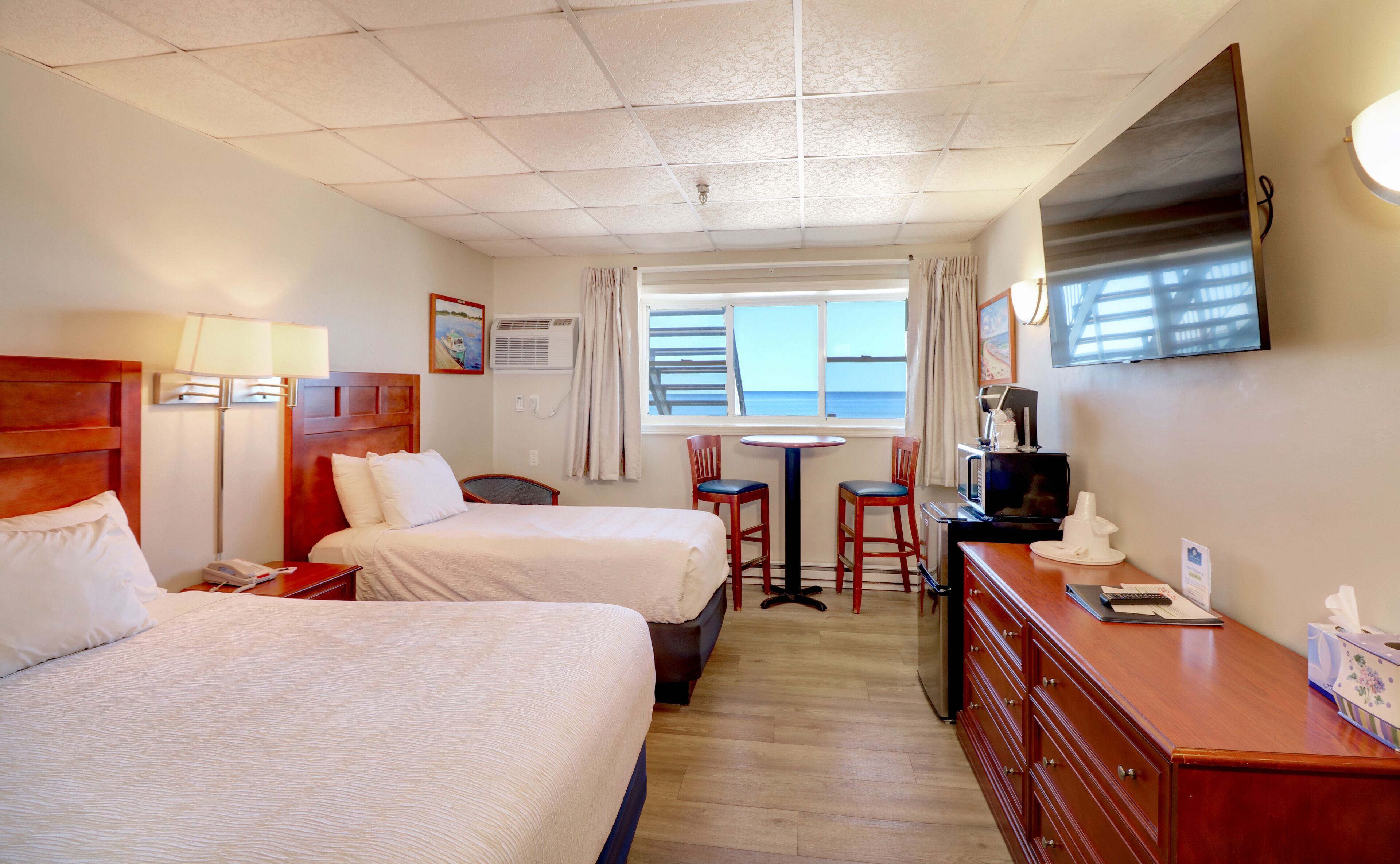 Ett hotellrum med två sängar, ett skrivbord med en tv, ett litet bord och ett fönster med utsikt över havet.