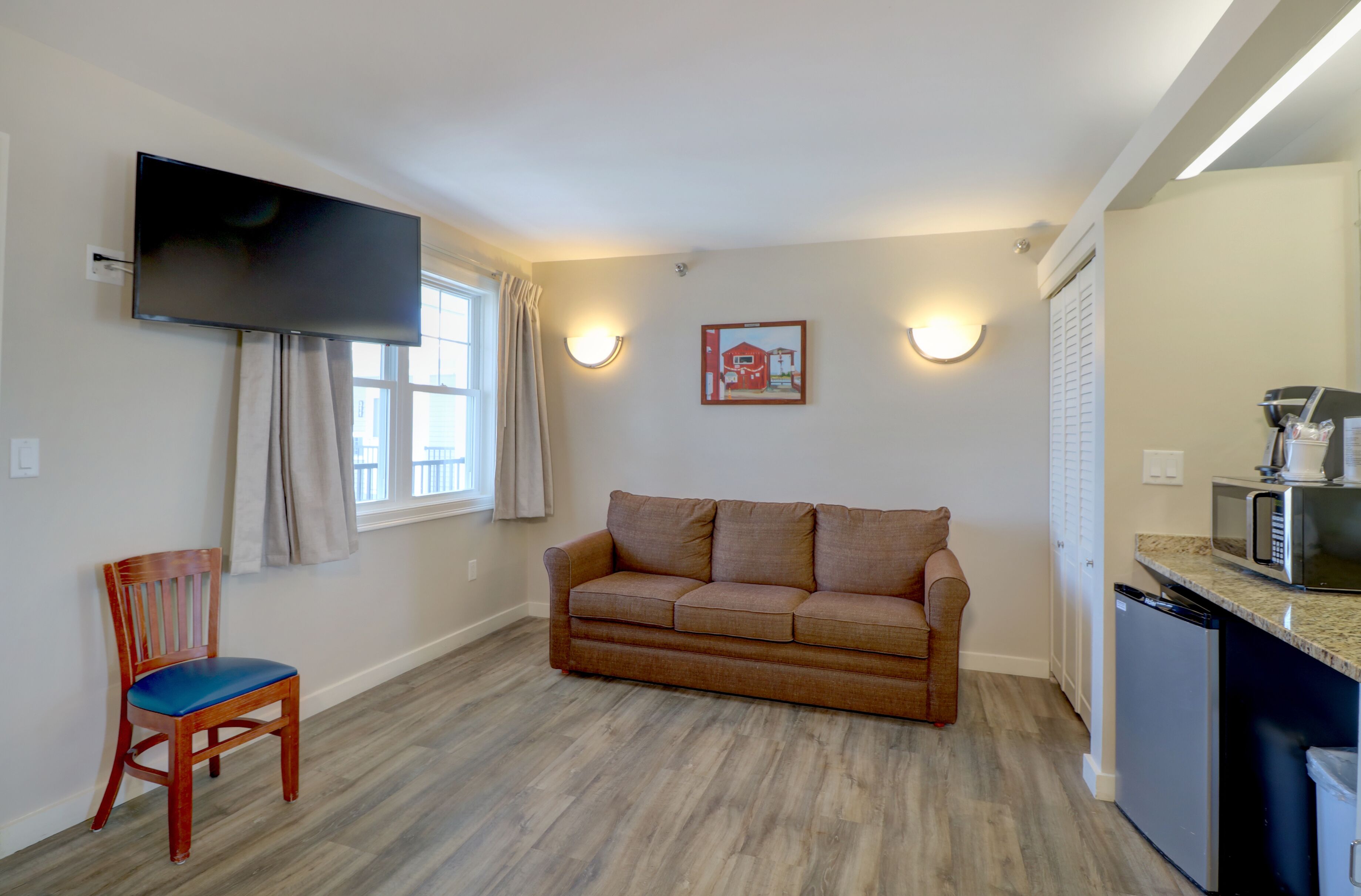 A living room with a sofa, a wooden chair, a flat-screen TV mounted on the wall, and a kitchen area with a microwave.