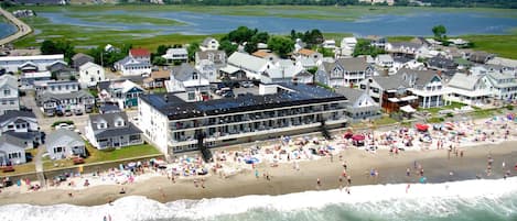 A coastal hotel with multiple floors, a beach in front, and a view of the sea and greenery in the background.
