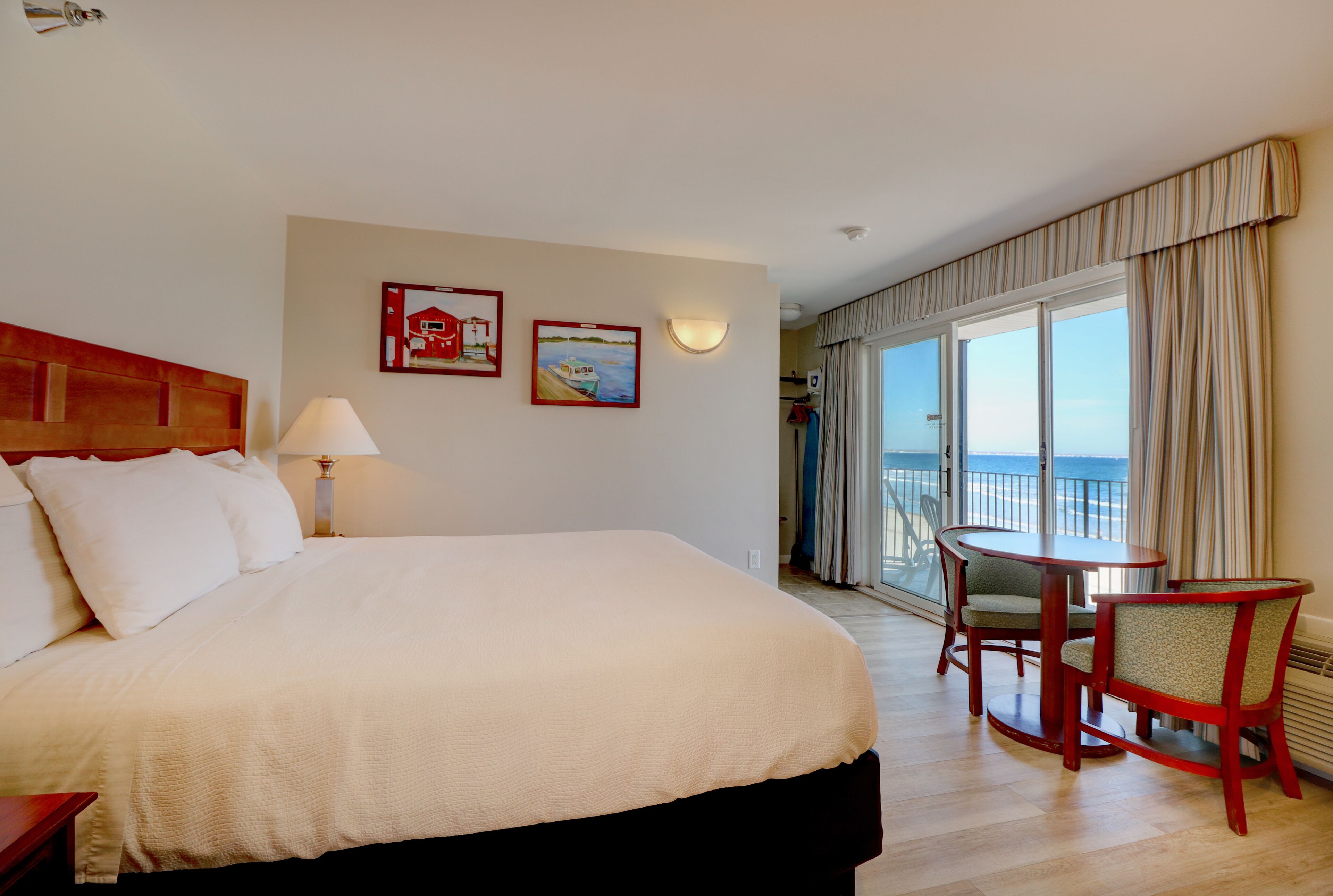A hotel room with a large bed, a desk with two chairs, a balcony with a view of the sea, and two framed pictures on the wall.