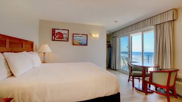 A hotel room with a large bed, a desk with two chairs, a balcony with a view of the sea, and two framed pictures on the wall.