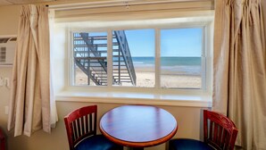 A room with a large window overlooking a beach and a staircase leading to the beach. There is a round wooden table with two chairs in front of the window.