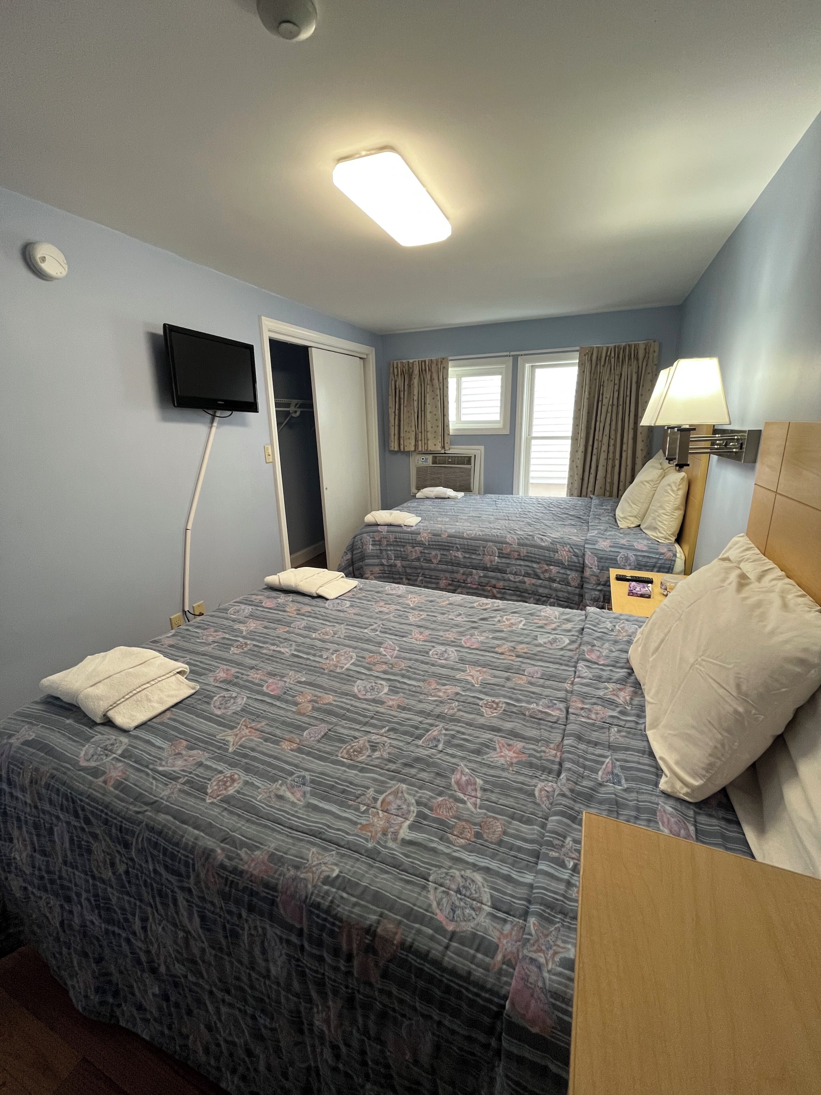 A hotel room with two beds, a TV mounted on the wall, a bedside lamp, and a window with curtains.
