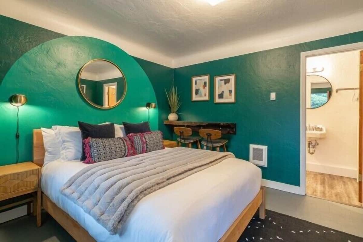 A bedroom with a bed, bedside tables, a mirror, wall art, and a door leading to a bathroom.