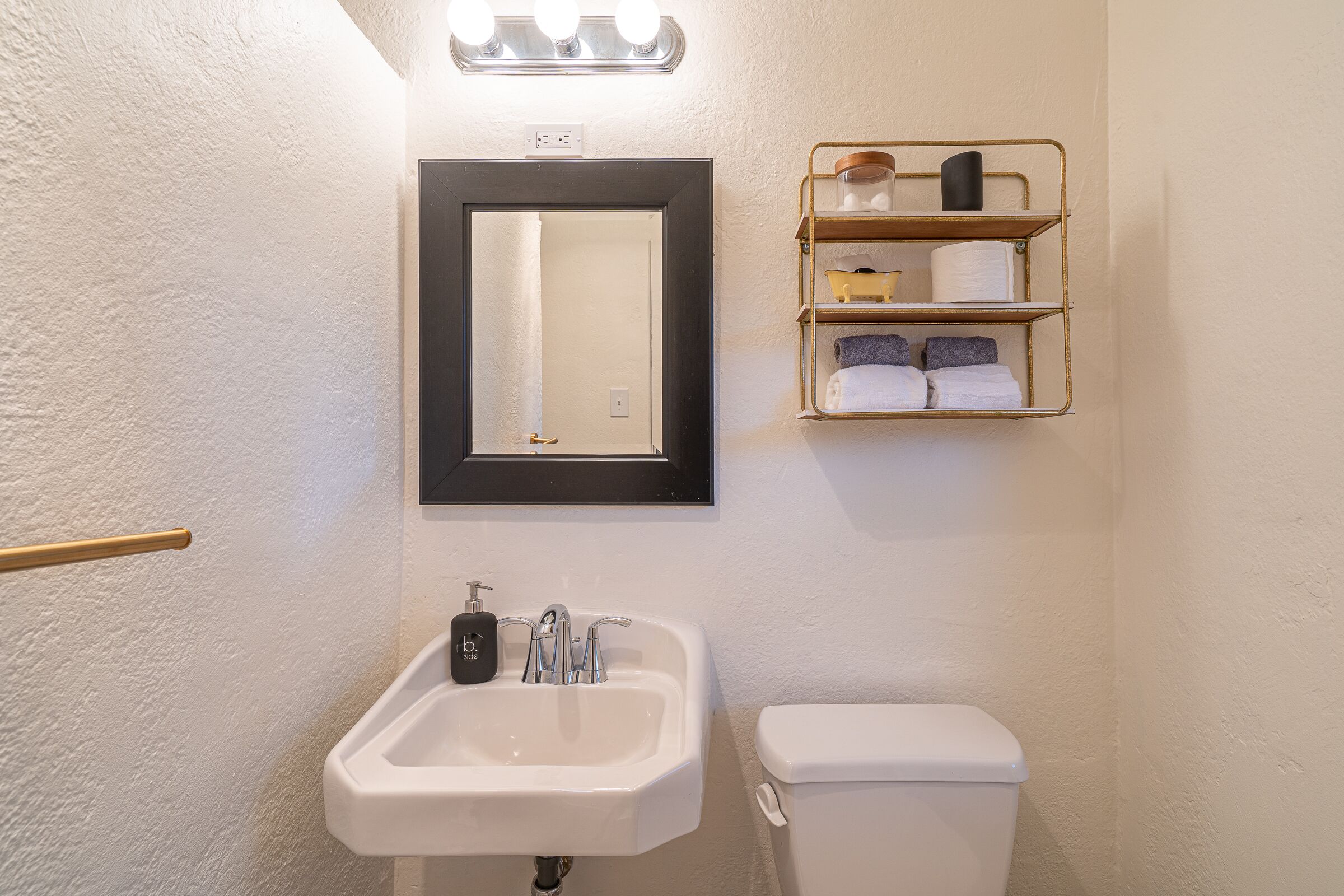 A bathroom with a white sink, a mirror, and a toilet. There is a shelf above the mirror with toiletries.