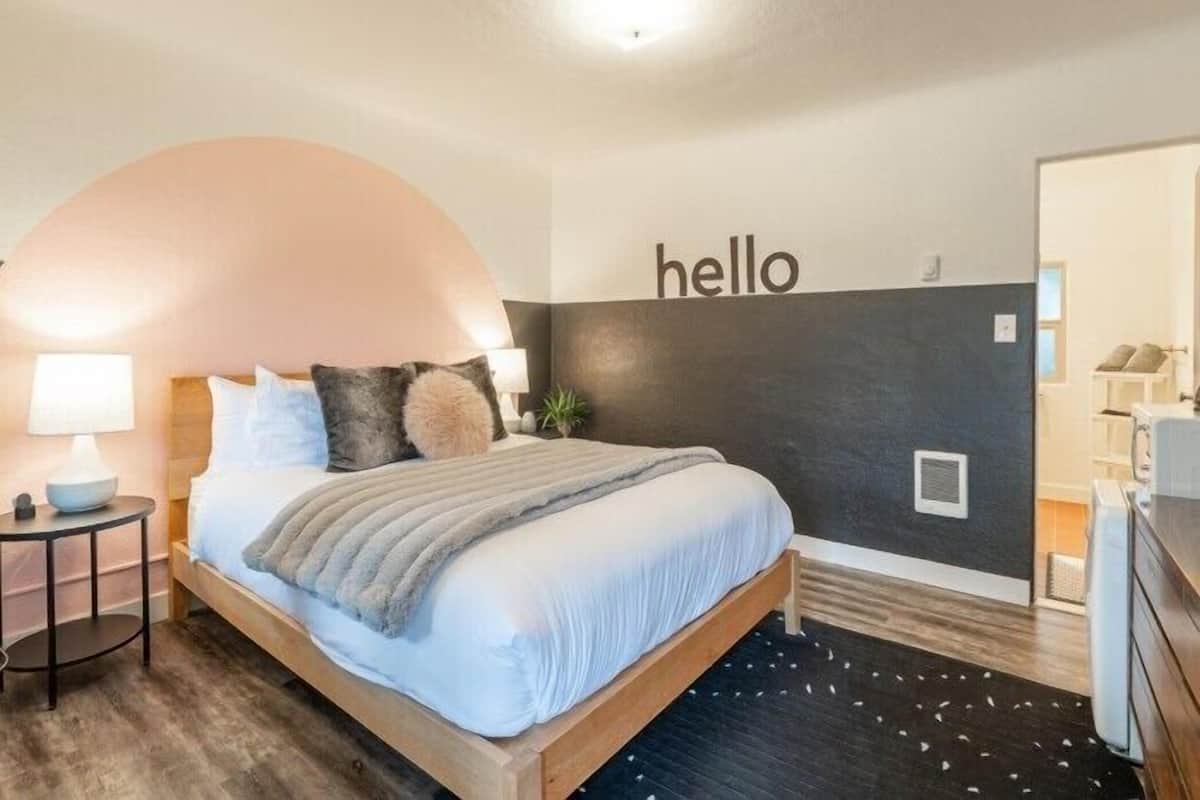 A modern bedroom with a bed, a nightstand, a lamp, a wall-mounted shelf, and a 'hello' sign.