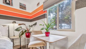 Una habitación moderna con una cama blanca, una mesita con una maceta roja y una ventana con vistas a árboles.