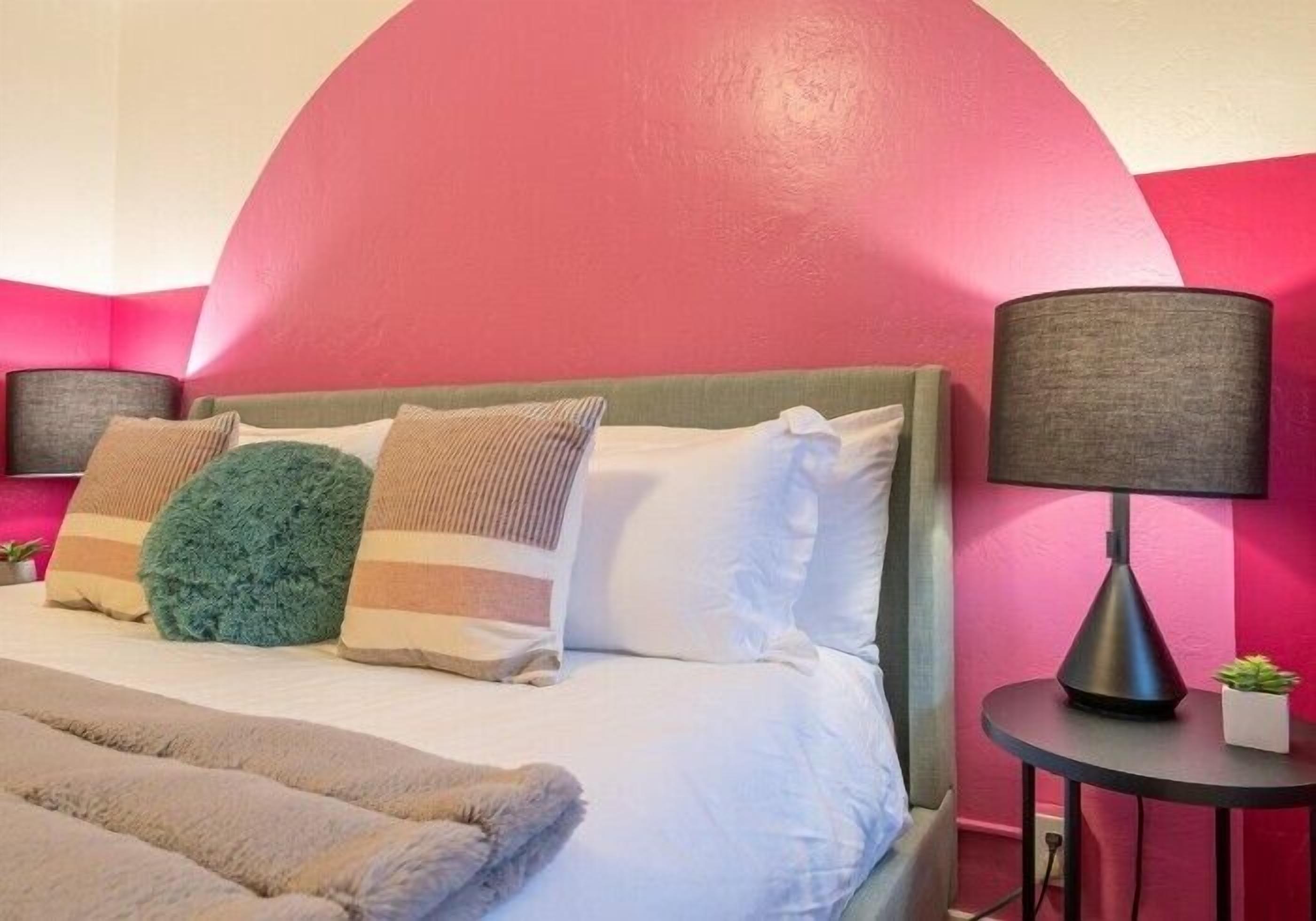 Una cama con varias almohadas, una lámpara de noche y una planta pequeña en una mesita.