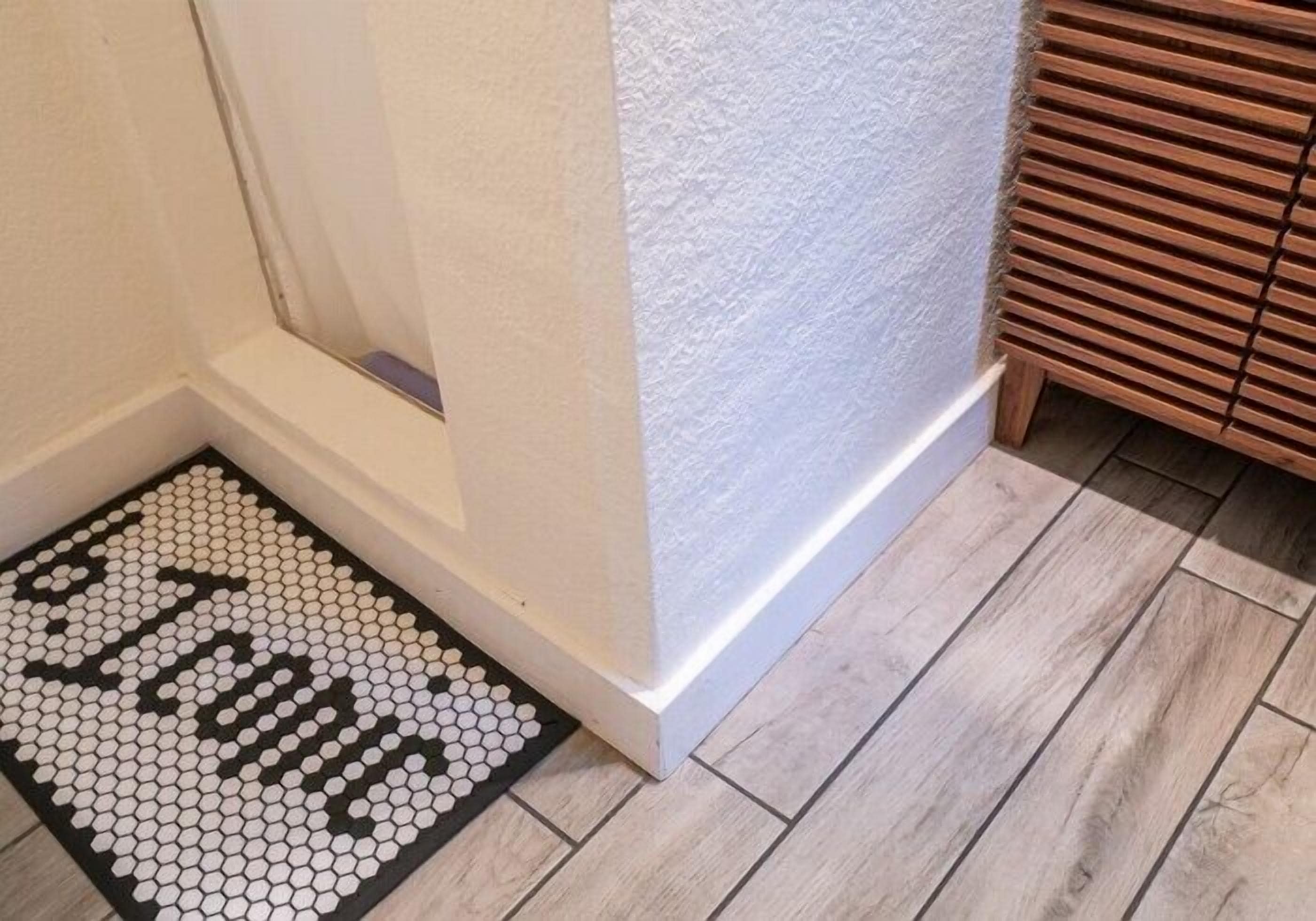 Un bagno con una parete bianca strutturata, pavimento in legno e tappetino d'ingresso in bianco e nero con piastrelle esagonali con la scritta "HOME".