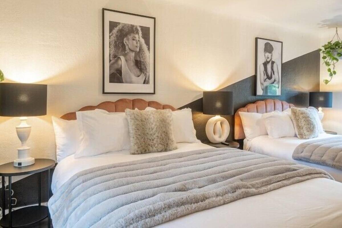 A hotel room with two beds, each with a fluffy pillow and a headboard. There are framed pictures on the wall, a bedside table with a lamp, and a plant hanging from the ceiling.