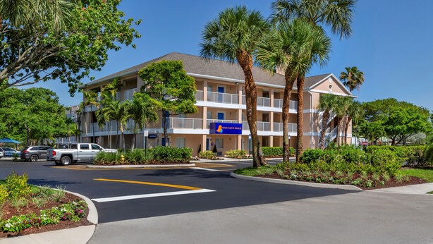 A multi-story hotel with a blue sign, palm trees, and a parking lot. - InTown Suites Extended Stay Fort Lauderdale (Tamarac)