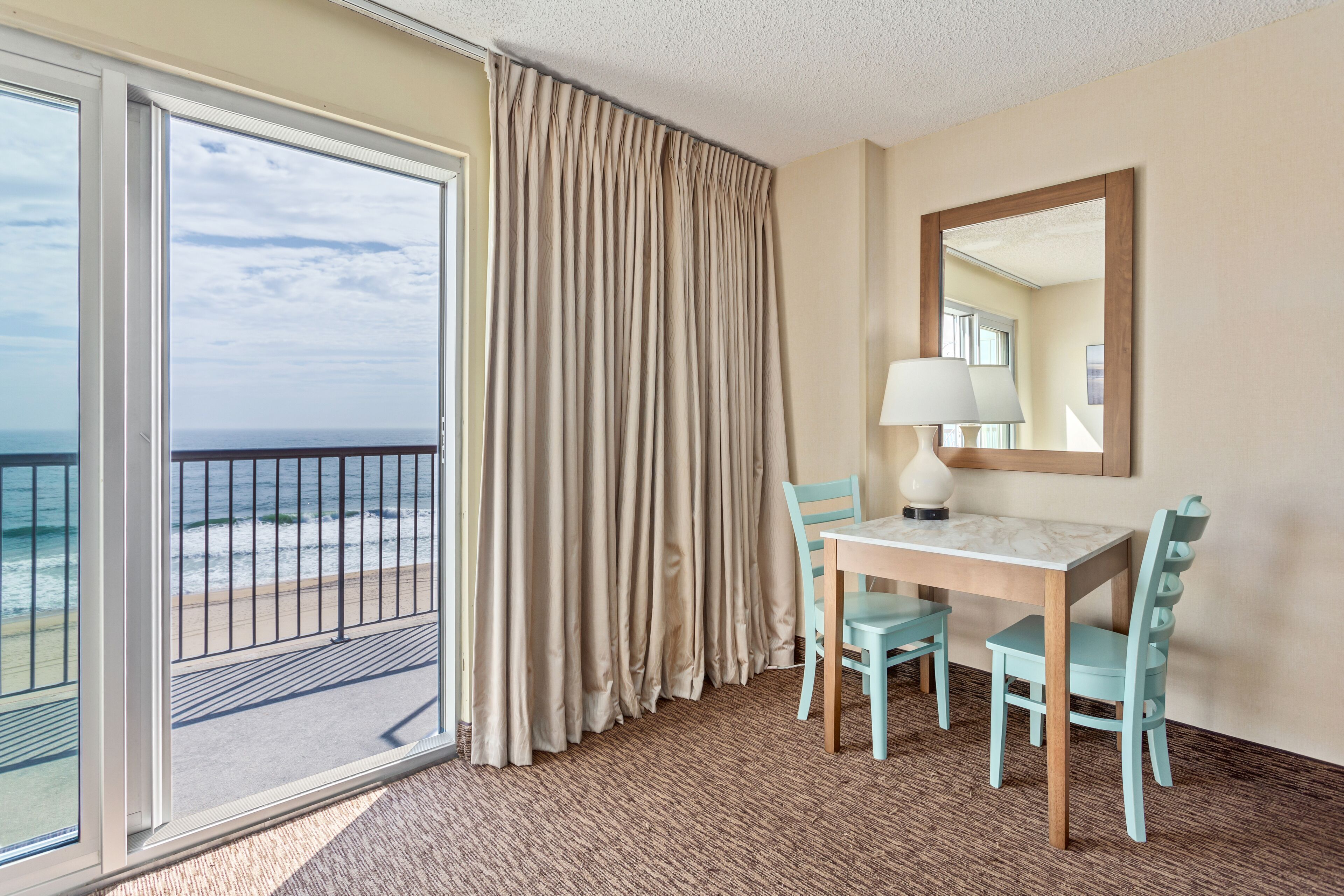 A hotel room with a balcony, a dining area with a table and chairs, a lamp, and a mirror on the wall.