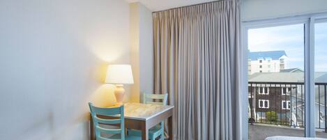 A hotel room with a bed, a desk with a lamp, two chairs, a large window with curtains, and a view of buildings outside.