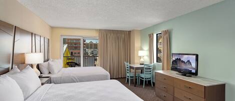 A hotel room with two beds, a desk with chairs, a TV, and a view of the outside.