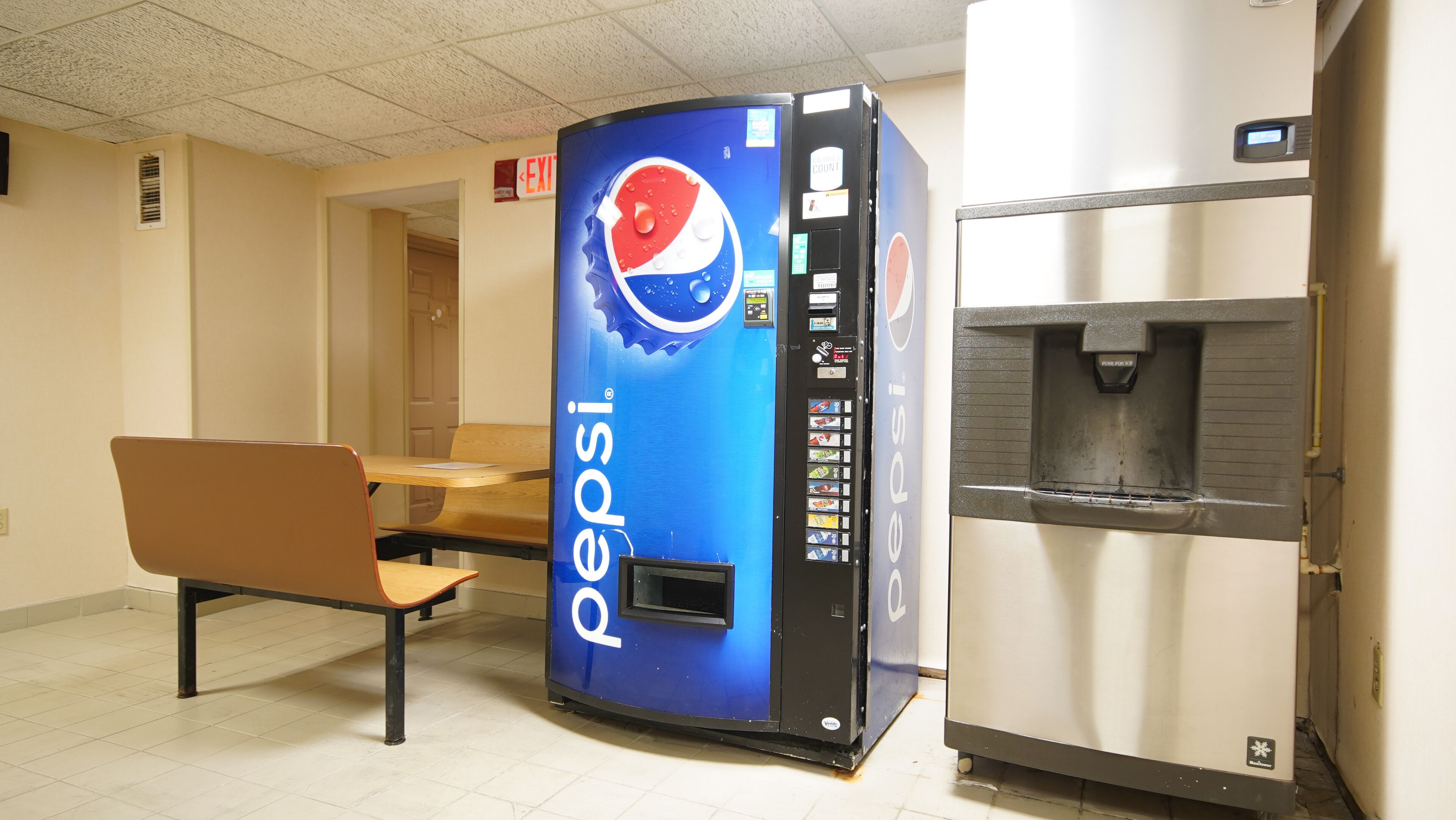 A vending machine with a Pepsi logo, a water dispenser, and a table with chairs in a room with tiled flooring.